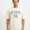 A male fashion model wearing an off-white oversized cotton t-shirt with a blue "HILFIGER" and athletic emblem chest print, paired with black trousers against a plain white background.
