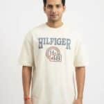 A male fashion model wearing an off-white oversized cotton t-shirt with a blue "HILFIGER" and athletic emblem chest print, paired with black trousers against a plain white background.