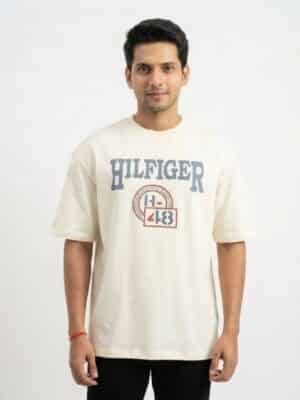 A male fashion model wearing an off-white oversized cotton t-shirt with a blue "HILFIGER" and athletic emblem chest print, paired with black trousers against a plain white background.