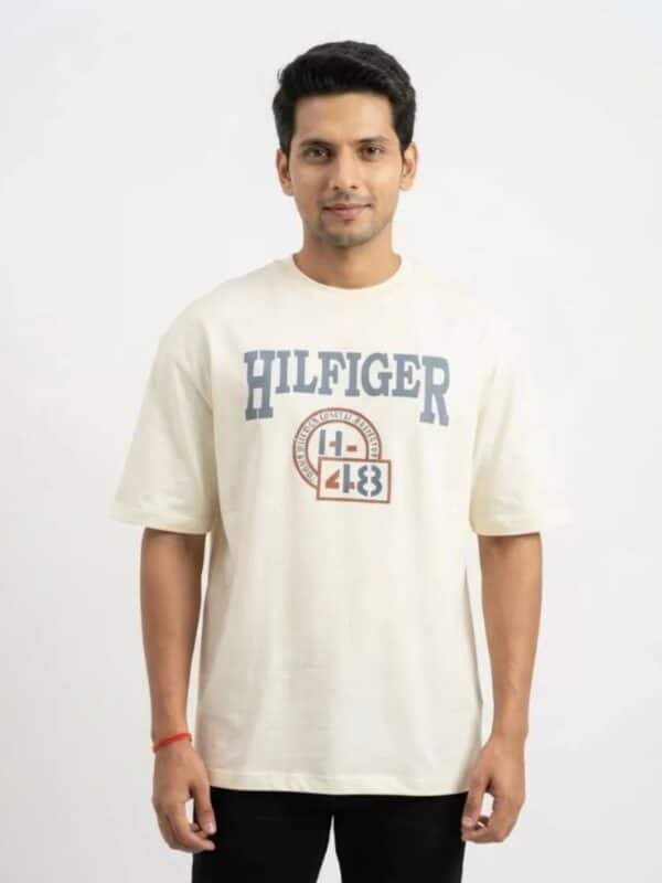A male fashion model wearing an off-white oversized cotton t-shirt with a blue "HILFIGER" and athletic emblem chest print, paired with black trousers against a plain white background.