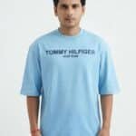 A male fashion model wearing a light blue oversized cotton t-shirt with a printed Tommy Hilfiger New Year chest logo and white trousers against a plain white background.
