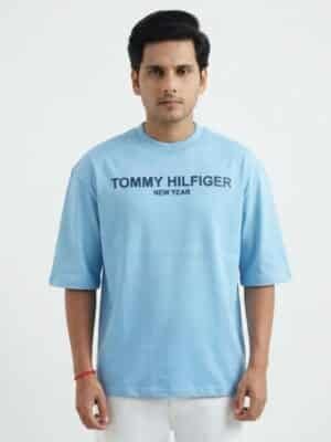 A male fashion model wearing a light blue oversized cotton t-shirt with a printed Tommy Hilfiger New Year chest logo and white trousers against a plain white background.