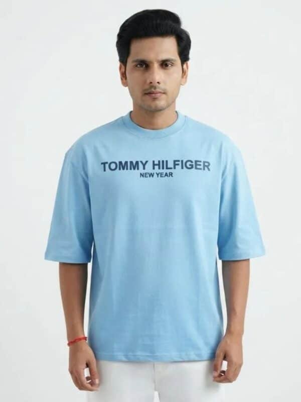 A male fashion model wearing a light blue oversized cotton t-shirt with a printed Tommy Hilfiger New Year chest logo and white trousers against a plain white background.