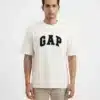A male fashion model wearing an off-white oversized cotton t-shirt with a bold black printed GAP logo on the chest and khaki trousers against a plain white background.