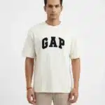 A male fashion model wearing an off-white oversized cotton t-shirt with a bold black printed GAP logo on the chest and khaki trousers against a plain white background.