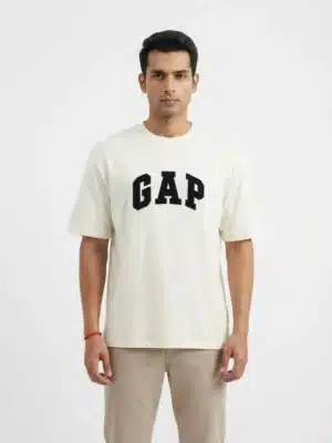 A male fashion model wearing an off-white oversized cotton t-shirt with a bold black printed GAP logo on the chest and khaki trousers against a plain white background.