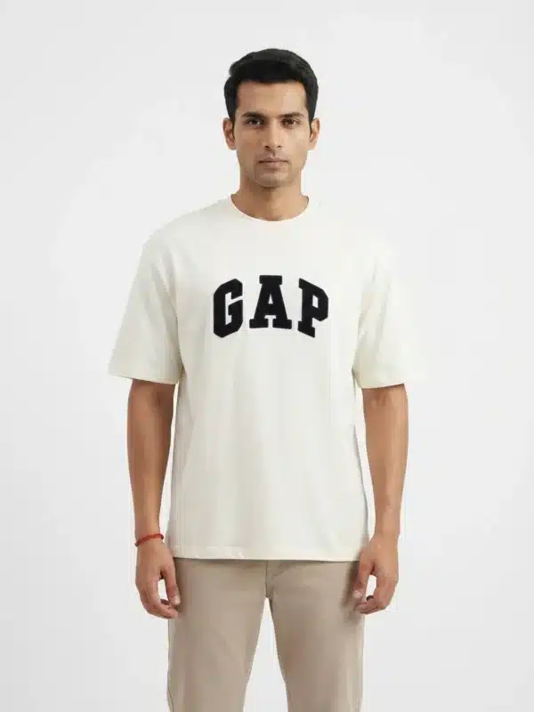 A male fashion model wearing an off-white oversized cotton t-shirt with a bold black printed GAP logo on the chest and khaki trousers against a plain white background.