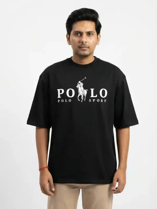 A male fashion model wearing a black oversized cotton t-shirt with a white printed polo sport chest logo and khaki trousers against a plain white background