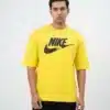 A male fashion model wearing a bright yellow oversized cotton t-shirt with a bold black printed Nike graphic logo on the chest and black cargo trousers against a plain white background.