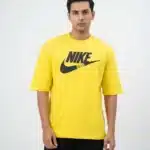 A male fashion model wearing a bright yellow oversized cotton t-shirt with a bold black printed Nike graphic logo on the chest and black cargo trousers against a plain white background.