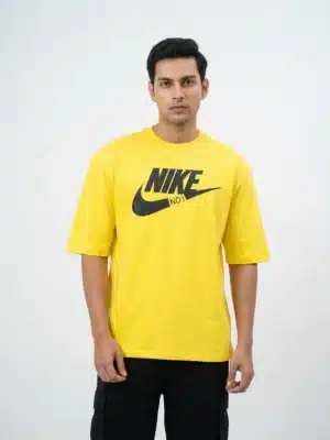 A male fashion model wearing a bright yellow oversized cotton t-shirt with a bold black printed Nike graphic logo on the chest and black cargo trousers against a plain white background.